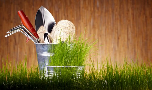 A close-up of a small garden scene on a wooden surface shows a metal container holding gardening tools including a small hand rake, a trowel, and a garden fork, alongside a ball of twine. In front of the container, there is a clump of fresh, green grass with thin blades, contrasting with the tools and extending slightly beyond the container's edge. The background is a warm, polished wooden surface, providing a natural and rustic ambiance suitable for a professional gardening context. This arrangement depicts essential gardening supplies in a tidy manner, reflecting readiness for outdoor gardening tasks typical in residential or small-scale garden maintenance, aligned with services offered by Gardening Services Spitalfields in the East London area.